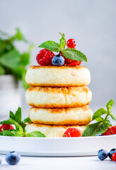 Thick pancakes with cottage cheese with blueberries, raspberries and red currants, decorated with mint on white plate, delicious breakfast. Gray table background
