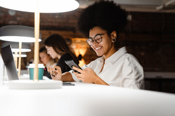 Smiling african woman using smartphone while working on laptop in office