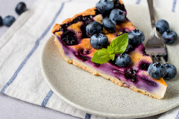 A piece of shortbread pie with blueberries with a mint leaf in a plate next to cutlery and a napkin, on a gray background, horizontal.