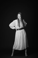 Young serious woman isolated on wall with copy space. Beautiful girl with hands on waist looking at camera. Portrait of lady in long dress looks confident on black background. Black and white photo.