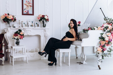 Pregnant woman strokes hugging the belly tummy abdomen enjoying pregnancy sitting grand piano dressed black suit. Future family, baby infant expecting child inside

