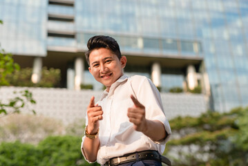 An assured young asian man makes a double finger gun gesture. Outdoor city scene.