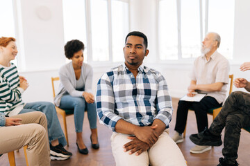 Portrait of sad African young man pensive looking away sitting in circle at group psychological counselling. Black male posing for picture treatment at therapy session. Concept of mental health