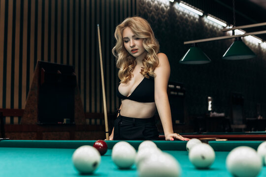 Blond woman playing enjoying billiard, hold cue, white billiard balls on table with green surface in billiard club. Pool game snooker pyramid player
