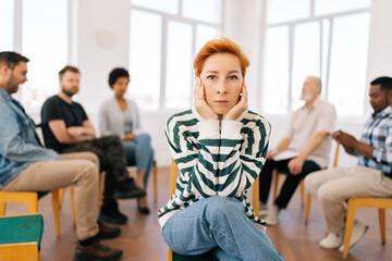 Portrait of depressed young woman covering ears with hands looking at camera sitting in circle at group psychological counseling. Concept of mental health, psychotherapy, social issues, support.