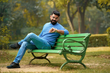 Young Indian man sitting at park and looking time in wristwatch.