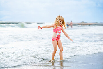 Happy Child, Little Preschool Girl in Swimmsuit Running And Jumping In The Waves During Summer Vacation On Exotic Tropical Beach. Family Journey On Ocean Coast.