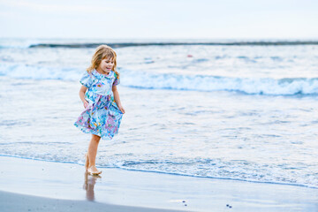 Fototapeta na wymiar Happy Child, Little Preschool Girl in Dress Running And Jumping In The Waves During Summer Vacation On Exotic Tropical Beach by Sunset. Family Journey On Ocean Coast.