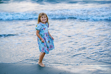 Happy Child, Little Preschool Girl in Dress Running And Jumping In The Waves During Summer Vacation On Exotic Tropical Beach by Sunset. Family Journey On Ocean Coast.