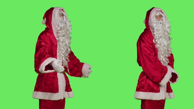 Adult in costume looks at wristwatch to check time against greenscreen backdrop, wearing santa claus red festive suit. Person acting like saint nick to celebrate winter holiday season.