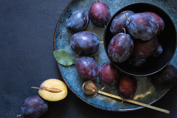 Fruits on the textured black table surface. Ripe juicy plums with leaves on a blue plate with copy...