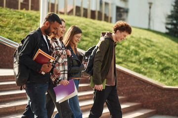 Walking, holding notepads. Four young students in casual clothes are together outdoors
