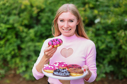girl in pink dress with long hair positively poses, holding fresh pink donuts with powder ready to enjoy sweets. 