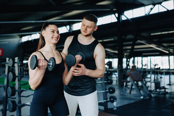 A dedicated male personal trainer works alongside a woman, providing assistance and instruction for her dumbbell lift in the gym. Fitness Progress Male Personal Trainer Assisting Woman in Gym Workout