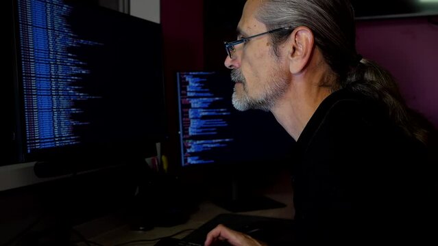Middle aged programmer with glasses and a ponytail coding in a darkened office