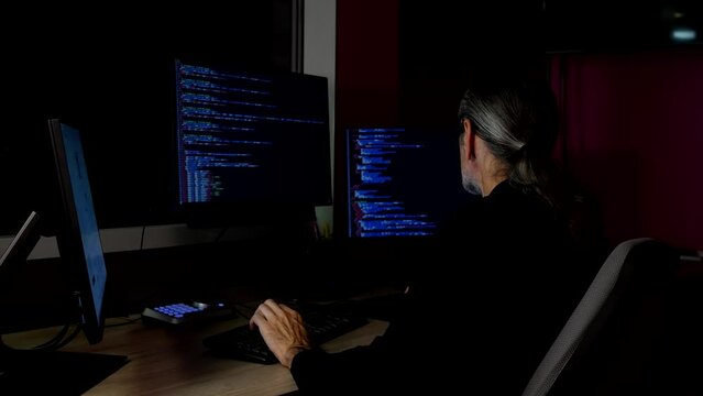 Middle-aged programmer with glasses and a ponytail and ponytail coding in a darkened office at a table with three monitors