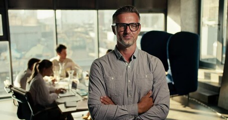 Successful middle-aged businessman in glasses and a blue shirt folded his arms on his chest, smiling and looking at the camera in an office with large windows