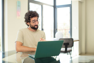 young adult bearded man with a laptop pointing at camera with a satisfied, confident, friendly smile, choosing you