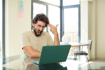young adult bearded man with a laptop looking unhappy and stressed, suicide gesture making gun sign...
