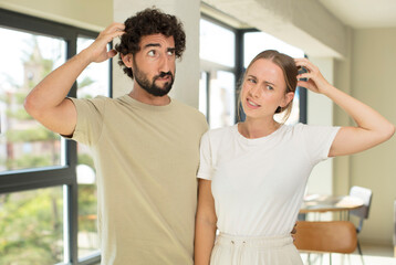 Fototapeta premium young adult couple feeling puzzled and confused, scratching head and looking to the side