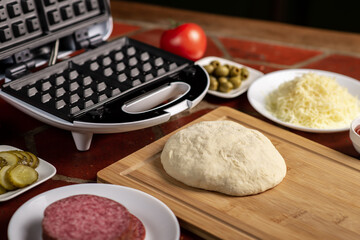 Cooking pizza waffle, piffle. Waffle iron and yeast dough for pizza and various ingredients - salami, cheese, olives, tomato on the kitchen table.