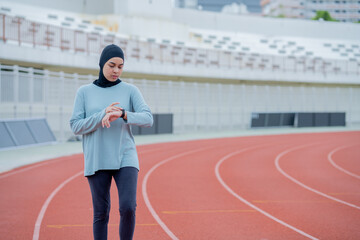 Fototapeta premium A young asian Muslim woman wearing a black hijab is running and checks progress on a smartwatch with a heart rate monitor at an outdoor stadium in the morning. Modern Muslim woman concept,Muslim sport