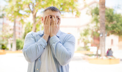 handsome man covering face with hands, peeking between fingers with surprised expression and looking to the side