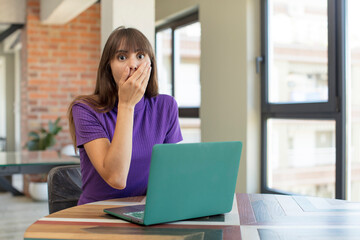 young pretty woman covering mouth with a hand and shocked or surprised expression.  desk laptop...