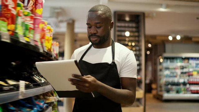 a Black-skinned man in a white t-shirt and a black apron walks along the windows of a supermarket and takes inventory using paper and a tablet