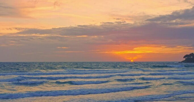 The sun is setting over the horizon and the waves of the sea are crashing towards the shore..Bright clouds as the sun sets over the horizon and waves of the sea come to shore..