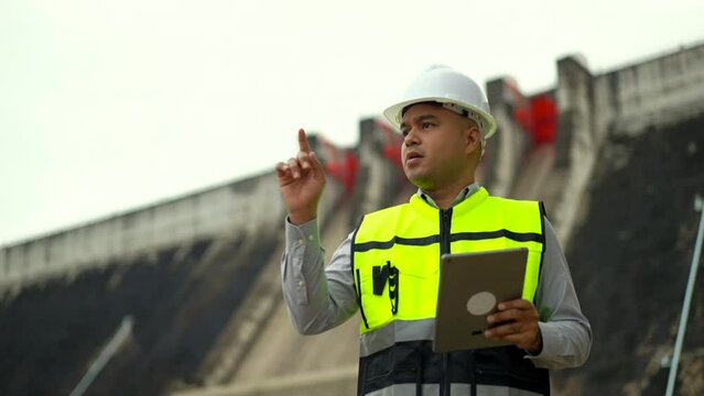 Professional asian maintenance engineer man inspection with tablet in construction site dam with hydroelectric power plant and irrigation. Engineer man wearing safety hat at project big building.