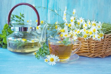 Tea from fresh chamomile flowers, mint, in a cup, on a wooden table, against the background of bouquets of fresh chamomile flowers