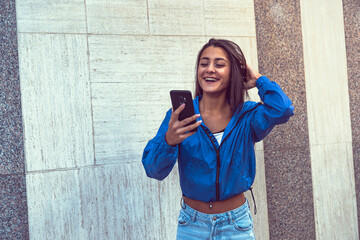 Stylish beautiful casual caucasian hip hop dancer chatting at smartphone in blue jacket, jeans, black top and white tshirt in urban town background. Outdoor city shooting of a model holding a phone