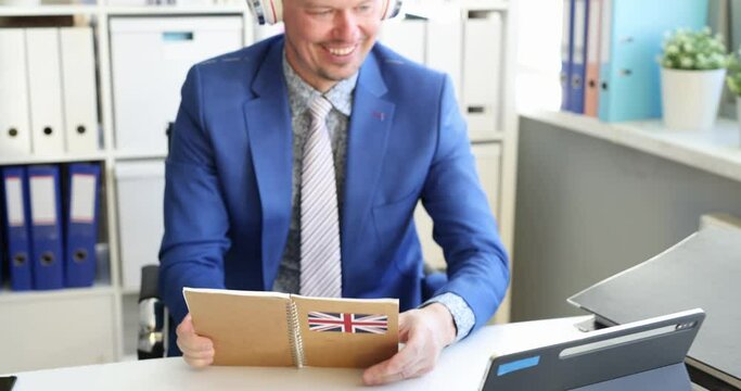 Young man with headphones holding english book in his hands and talking on computer video link 4k movie. Online foreign language courses concept