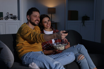 Happy couple watching TV at home in evening. Man changing channels with remote control