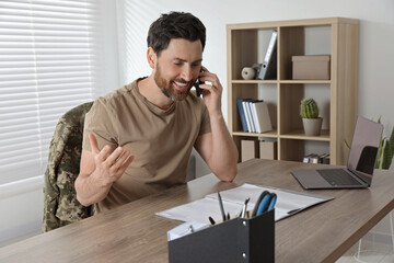 Happy soldier talking on phone at wooden table indoors. Military service