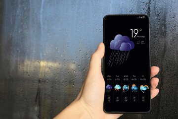 Woman checking weather using app on smartphone near wet window, closeup. Data, cloud with rain and...