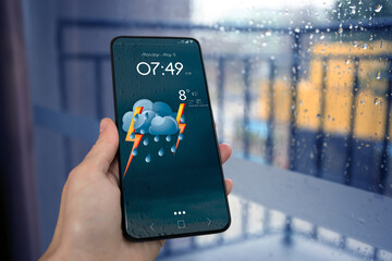 Woman checking weather using app on smartphone near wet window, closeup. Data and illustration of cloud with lightning and rain on screen - obrazy, fototapety, plakaty