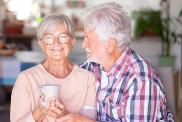 Senior white-haired man embracing and looking with affection at his wife drinking a cup of coffee or tea. Forever love, elderly couple in love