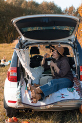 Young Happy woman relax at Car Trunk having breakfast with coffe and croissant. Vacation and Travel Concept. Vertical