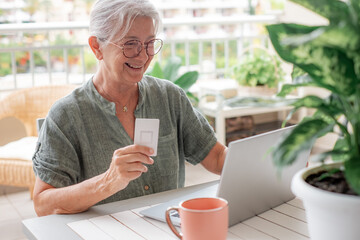 Online shopping concept. Happy senior woman using laptop sitting outdoors on terrace doing e-commerce shopping, senior woman in glasses holding credit card using modern technology