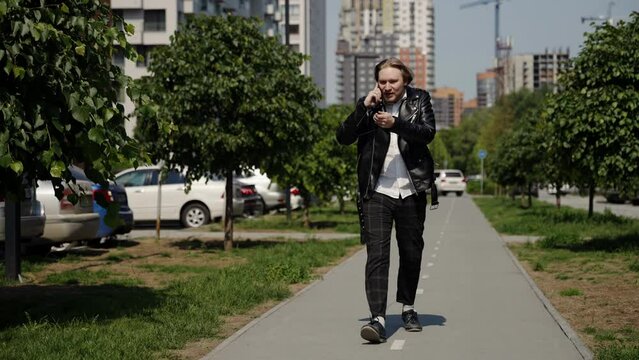 Scandal on the phone. Aggressive gestures in a young man who walks through a city block. Slow motion, summer mood, green trees, leather jacket.