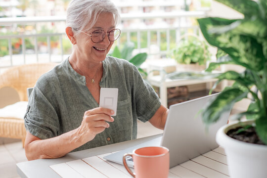 Online shopping concept. Happy senior woman using laptop sitting outdoors on terrace doing e-commerce shopping, senior woman in glasses holding credit card using modern technology