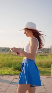 Vertical Video, Happy Woman with Slim Figure Smiles and Walks in City Park Using Mobile Phone for Social Networks. Beautiful Girl in White Hat Walks around Summer City Using Smartphone. Slow Motion.