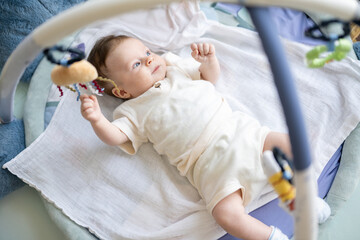 Top view of adorable little baby lying on children rug looking away and playing with toys.