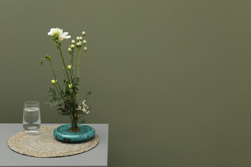 Stylish ikebana with beautiful flowers, green branches and glass of water on gray table near olive wall, space for text
