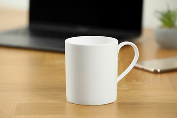 White ceramic mug on wooden table at workplace. Mockup for design