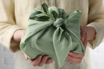 Fototapeta na wymiar Furoshiki technique. Woman holding gift packed in green fabric and decorated with ruscus branch, closeup