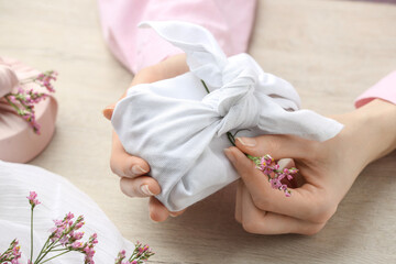 Furoshiki technique. Woman decorating gift wrapped in white fabric with beautiful pink flower at wooden table, closeup