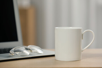 White ceramic mug, glasses and laptop on wooden table at workplace. Space for text